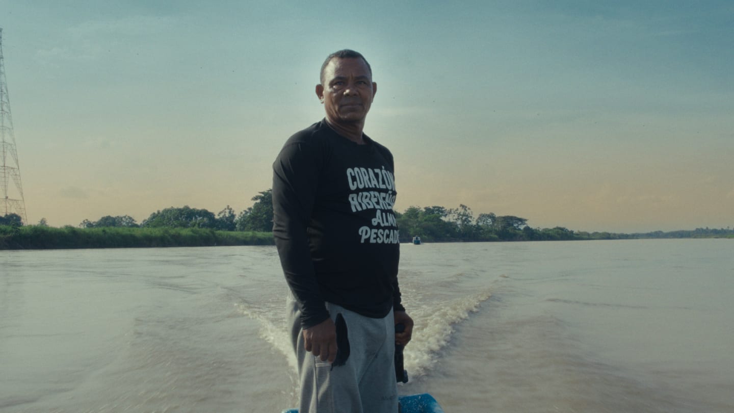 A man in a black shirt looks forward as he stands on a moving boat on a wide river with lush green banks.
