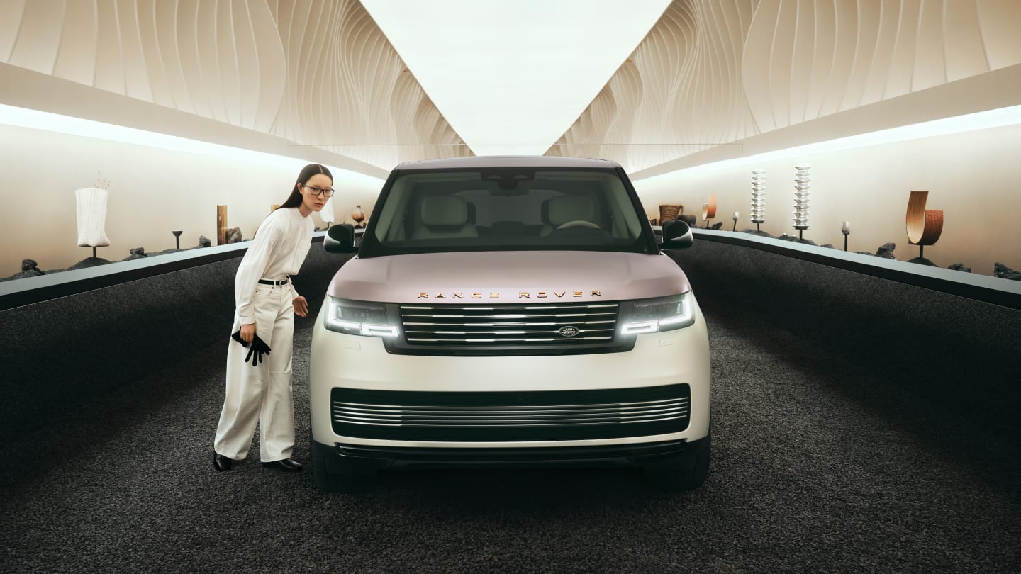 A woman in a light-colored outfit stands beside a two-tone white Range Rover, which is parked in a brightly lit, minimalist tunnel. Abstract art pieces are displayed on pedestals along the tunnel walls.
