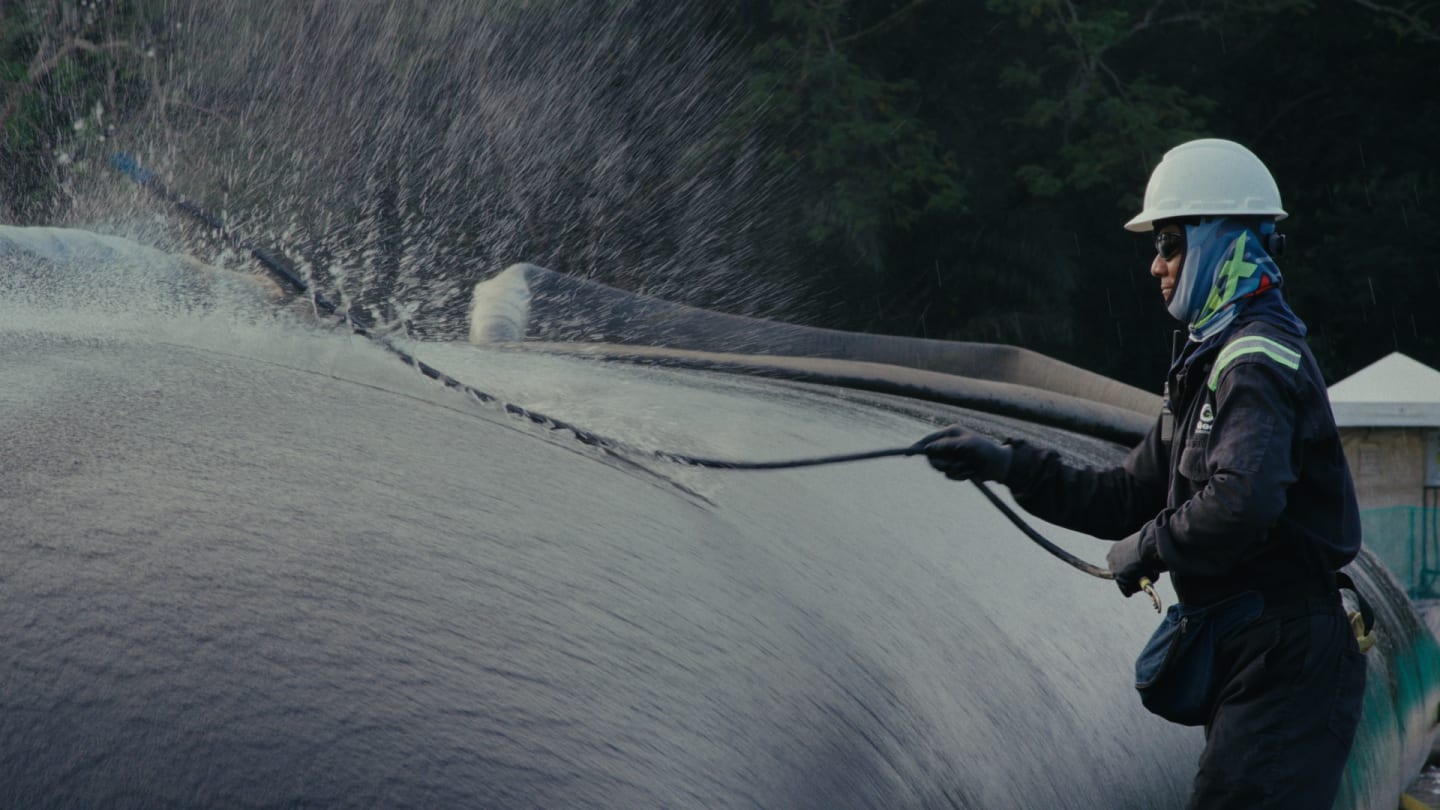 A worker in a hard hat and safety gear powerfully sprays water onto a large industrial surface, creating a fine mist.