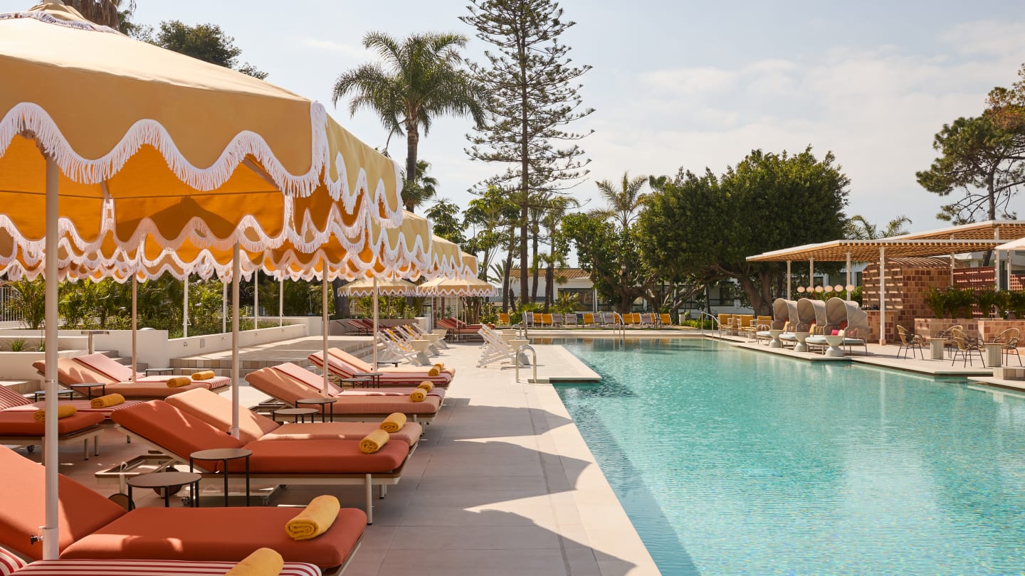 A sunny, luxurious outdoor resort pool area featuring a long, clear blue swimming pool. Alongside the pool are rows of orange-and-white striped lounge chairs shaded by retro-style yellow umbrellas with white scalloped trim. Lush green palm trees and foliage are visible in the background under a bright, clear sky.