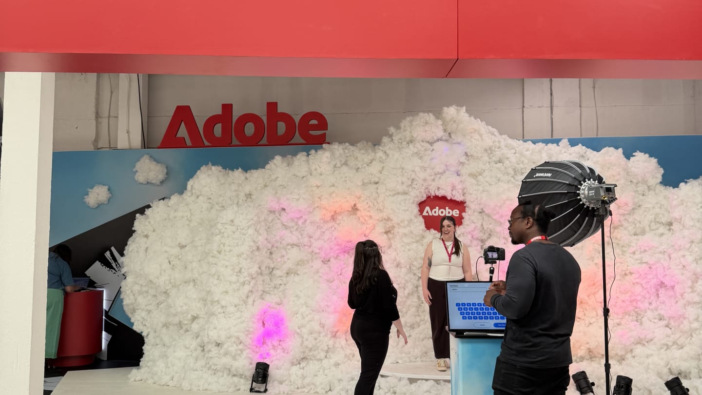 A presentation stage featuring an Adobe logo and a large backdrop resembling white clouds, with staff and audio-visual equipment in the foreground.