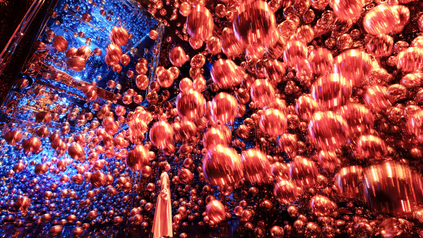 A person in a long light-colored outfit stands in a dark, highly reflective room filled with countless shimmering, copper and rose-gold spheres that appear to float freely in the air. The mirrored floor creates a perfect reflection below, and vibrant blue lighting glows softly in the background.