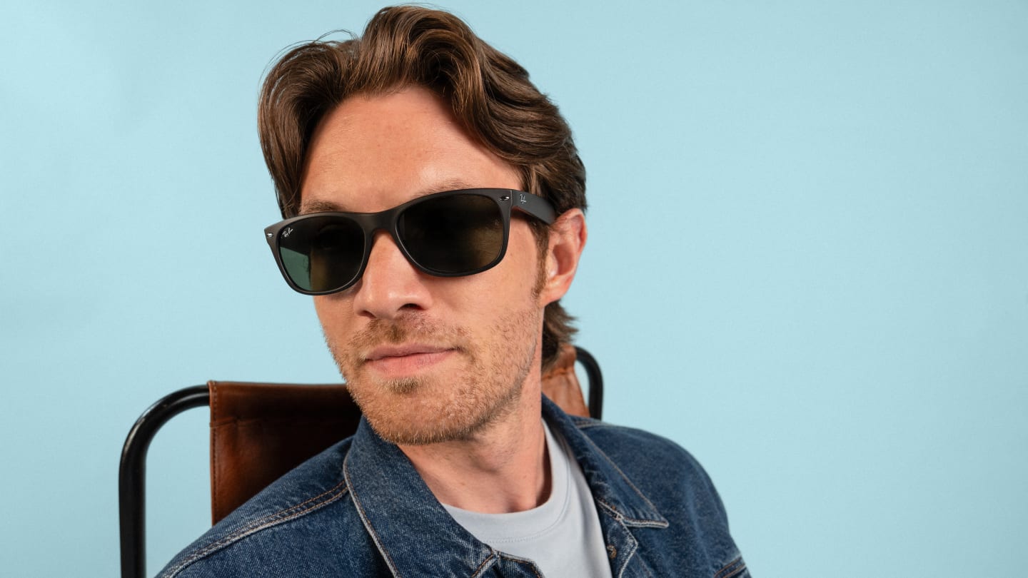 A man wearing black sunglasses and a denim jacket sits confidently against a light blue background.