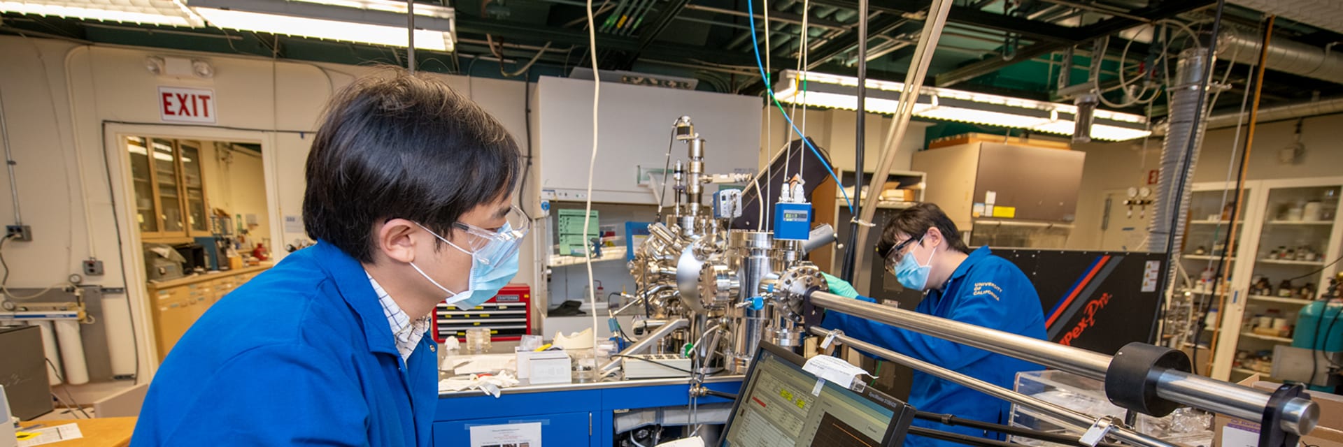 Two masked men wearing blue jackets and protective glasses and stand at opposite desks in a laboratory. The man in the forefront works on a desktop computer, and the man in the rear is handling metal machinery.