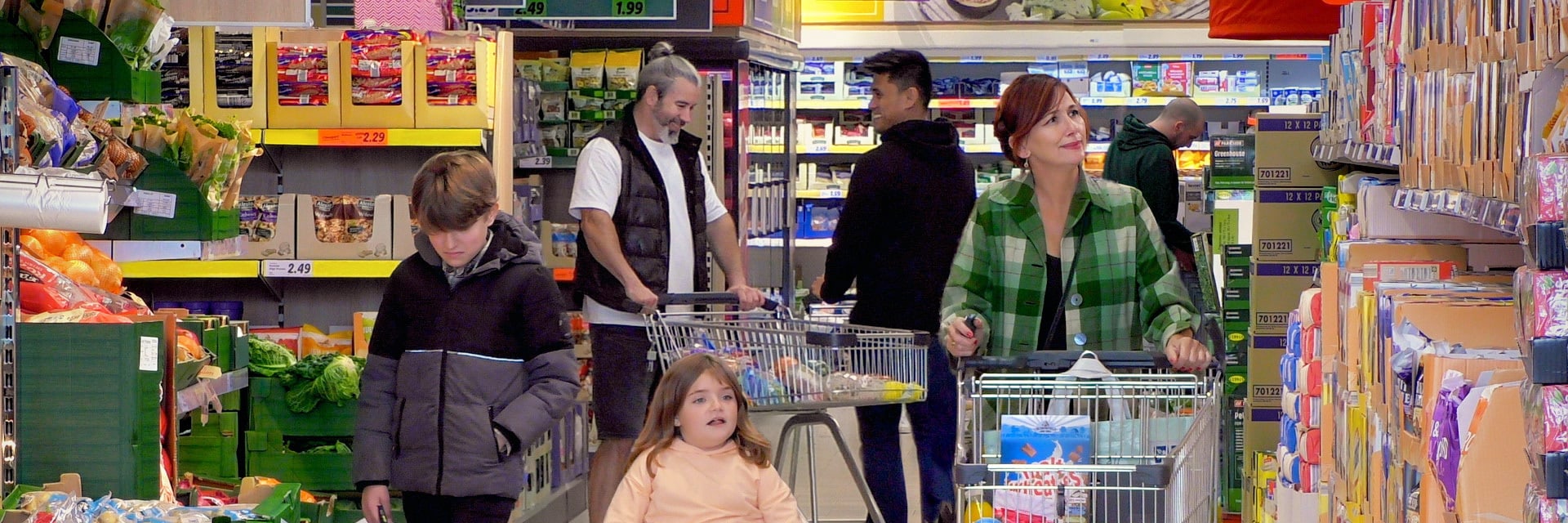 A mother pushes a trolley of shopping down the cereal aisle of a Lidl supermarket, with her teenage son and young daughter in a wheelchair beside her and other customers in the background