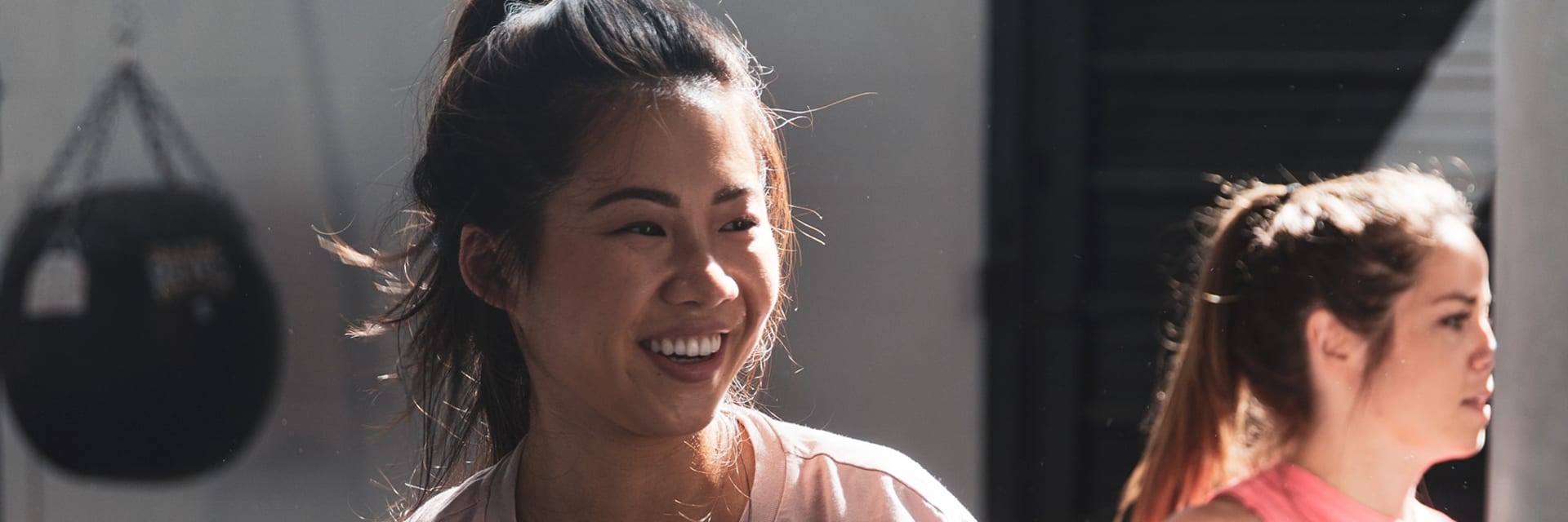 A woman with light skin and dark hair smiles as she works out. Her hair is in a high pony tail and she wears a pink T-shirt.