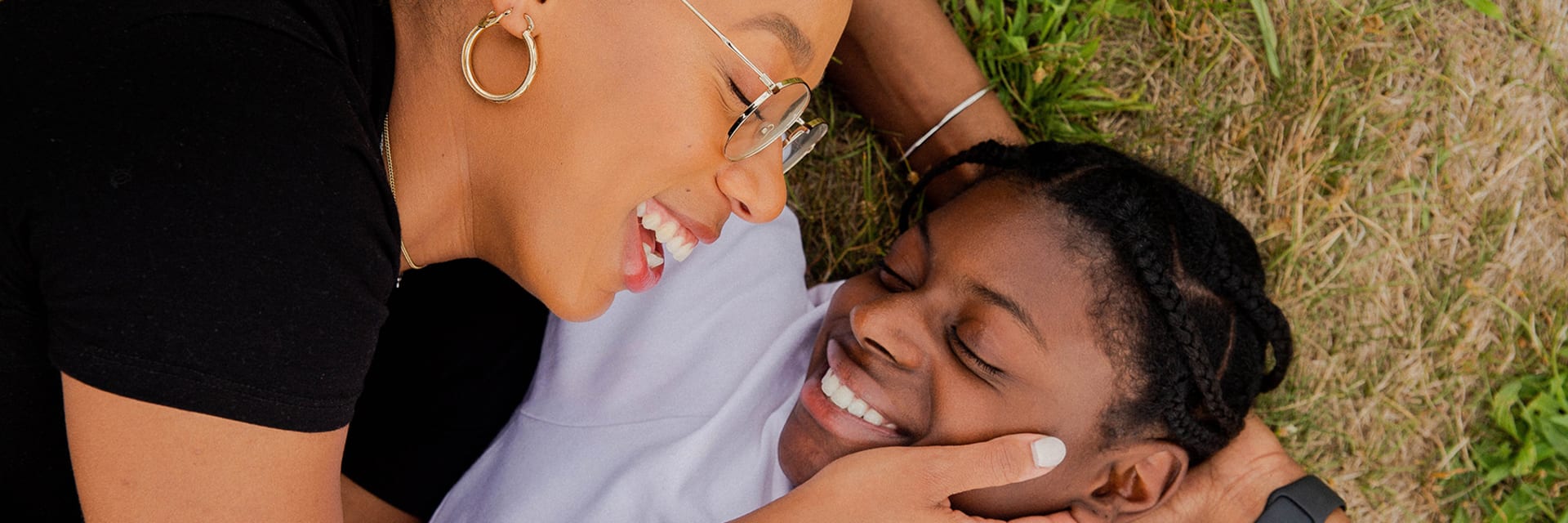 Two black women, smiling and laughing as they lay in the grass. On the left, a woman with gold-rimmed glasses and gold hoop earrings caresses the other woman's face, who lays with hands behind her head. Her hair is braided and she wears a white T-shirt.