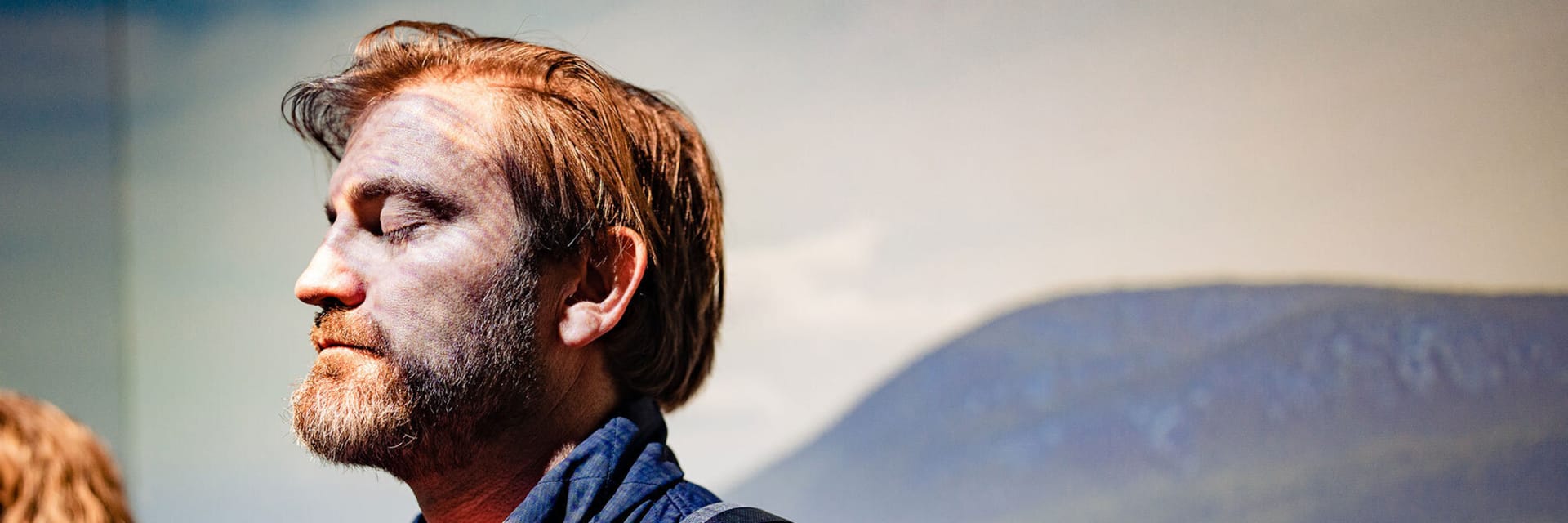 A white man with brown hair and a beard has his eyes closed and faces the viewer's left as a light shines on him. A mountainside backdrop rests behind him.