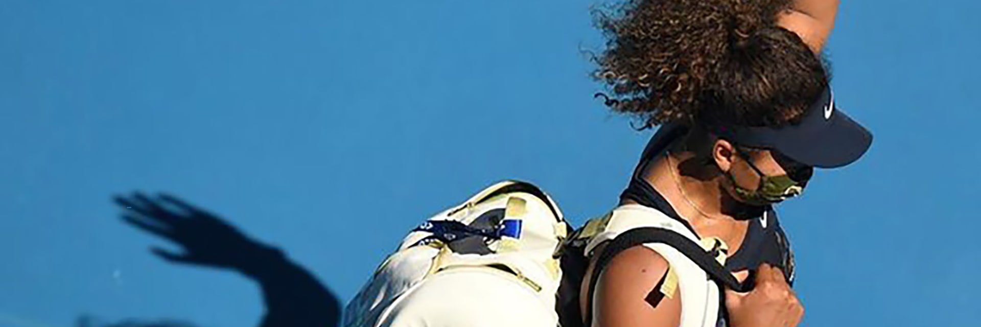 A woman with tan skin and dark, curly hair in a pony tail wears a black visor and waves above her head as she walks off-camera toward the viewer's bottom right.