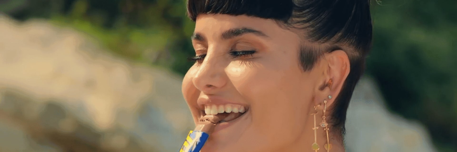 Girl with dark hair in a bun and long gold earrings smiling with her eyes closed as she eats a chocolate bar
