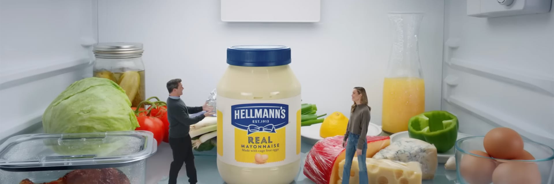 A man with dark hair and light skin stands opposite a woman with blonde hair and light skin. They are miniaturized and standing on a refrigerator shelf next to various produce items: an old chicken wing, lettuce, tomatoes, cheese, orange juice, eggs.