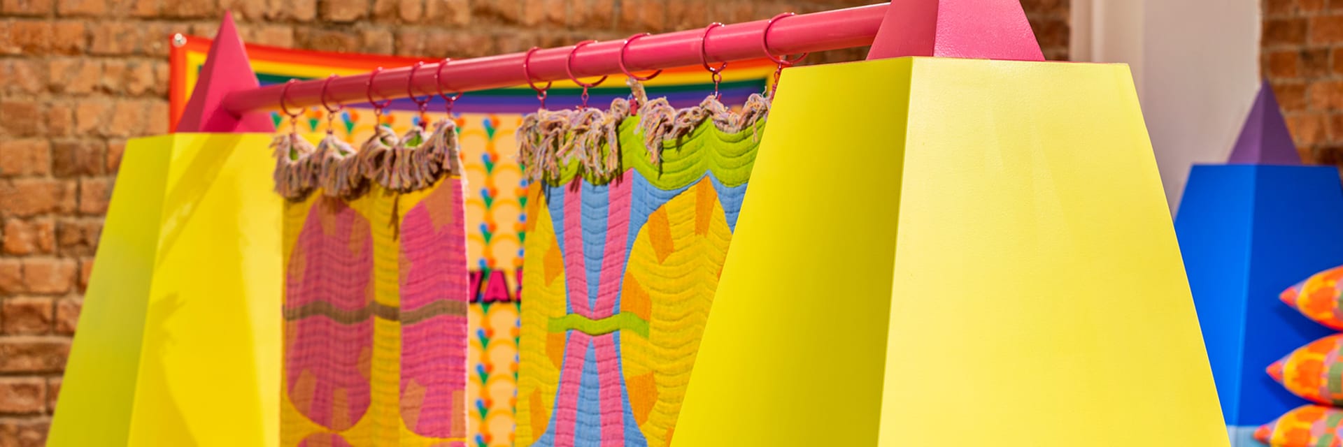 Two yellow pyramid pillars with pink tips hold a colorful tapestry that hangs between them. The image is angled with the right pyramid closer to the viewer.