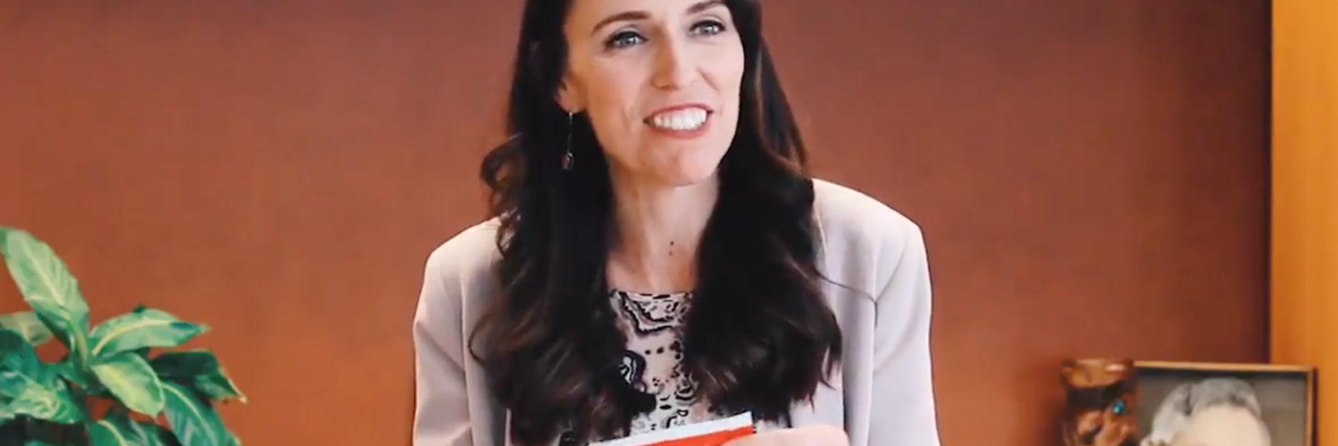 A woman with light skin and brown hair smiles to the right of the camera, holding an envelope. She is wearing a white blouse.