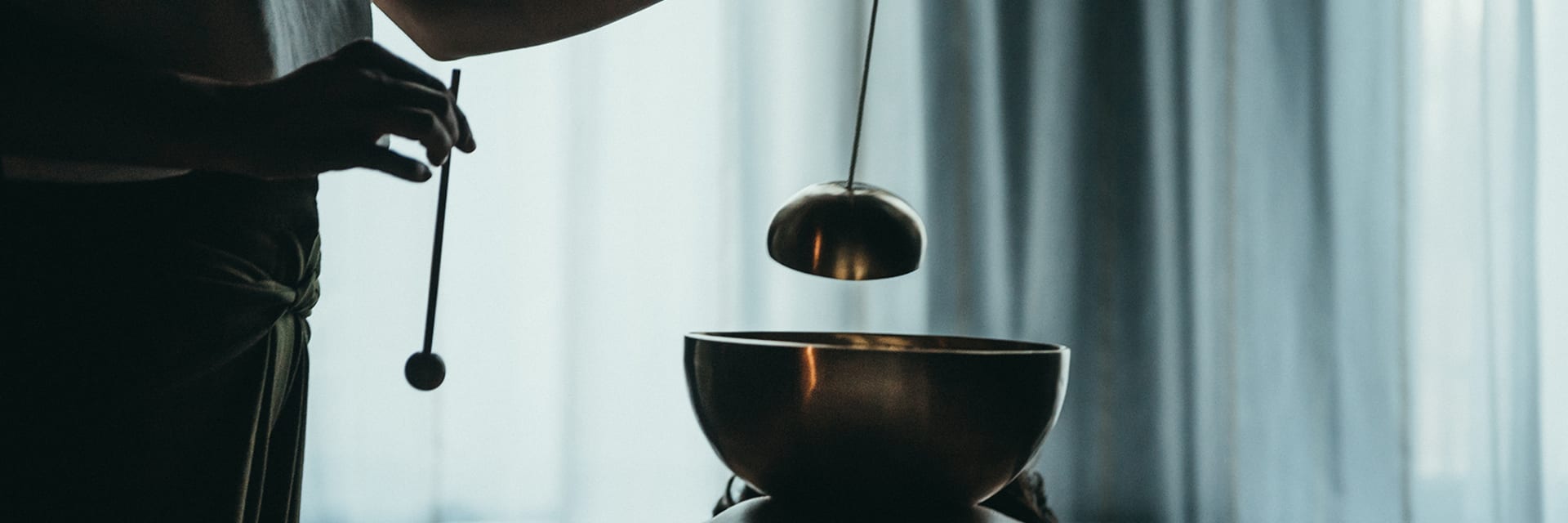 A person holds a chain with a ball at the end over a copper sonic bowl, which rests on a person's back.