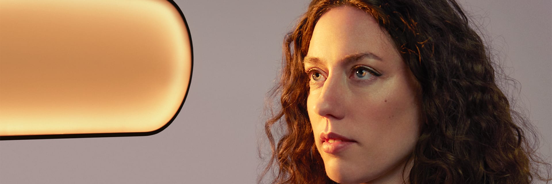 A woman with light skin and curly, long auburn hair is staring to the left at a pill-shaped light fixture.