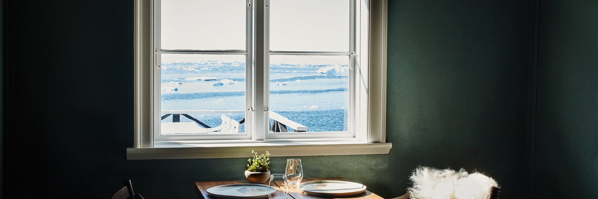 A table is set in front of a window in a room with dark green walls and two chairs. Through the window is an arctic ocean scene.