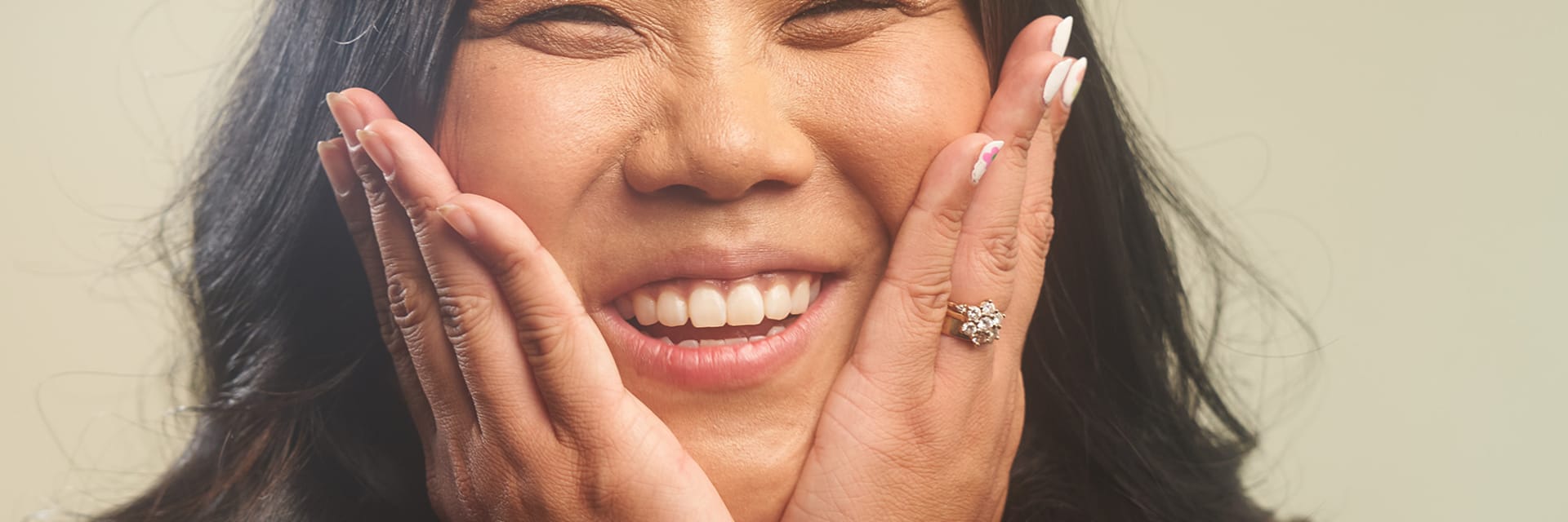 A woman with tan skin and brown hair smiles and holds her hands to either side of her face.
