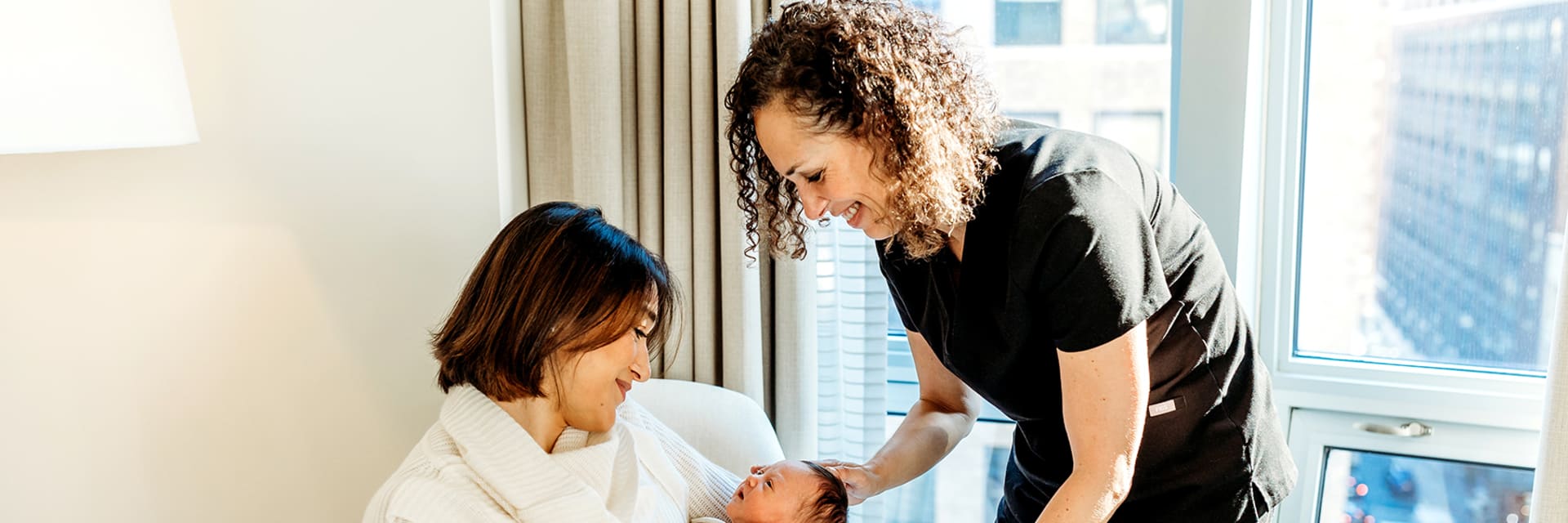A woman with short dark hair sits and smiles, holding a newborn baby with light skin and dark hair. A woman with light skin and curly, mid length hair smiles and stands next to them and leans over the baby.