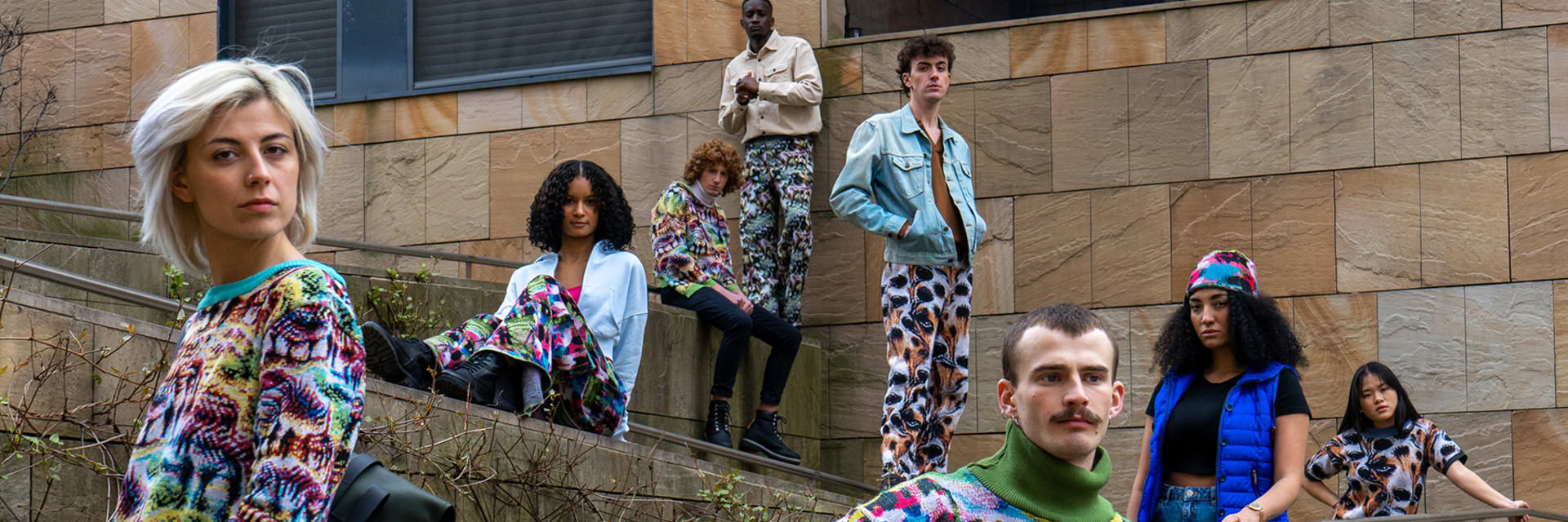 Models stand in an urban space with stone walls wearing similar, colorful animal-print patterned sweaters. A man and a woman stand in the forefront, and 6 models stand behind them on steps.