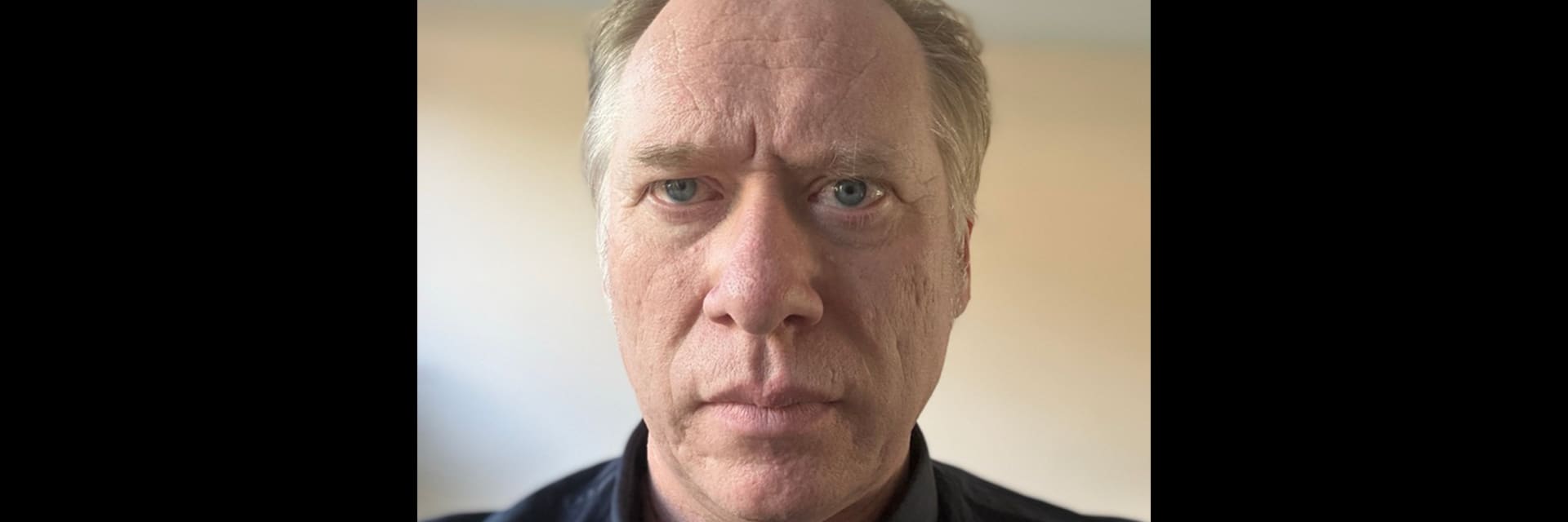 A white man with blue eyes and light blonde hair looks blankly into the camera. He wears a navy collared shirt.