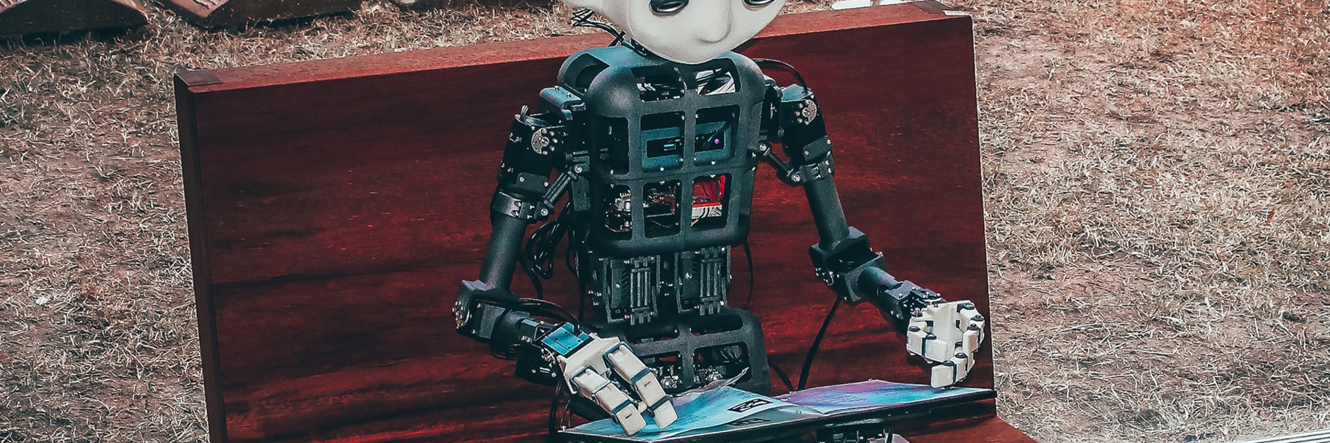 A digitally rendered avatar with a white robotic head and face sits on a red bench and flips the blue and purple pages of a magazine.