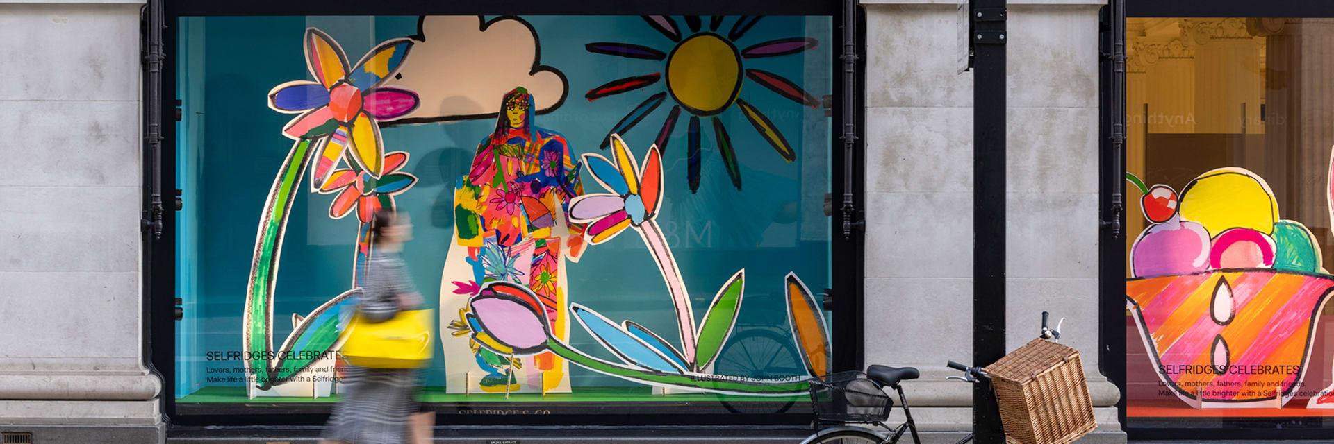 Window display on street with a teal background and bright, giant, colorful flowers with a sun in the corner. A woman walks by the display, blurred in movement.