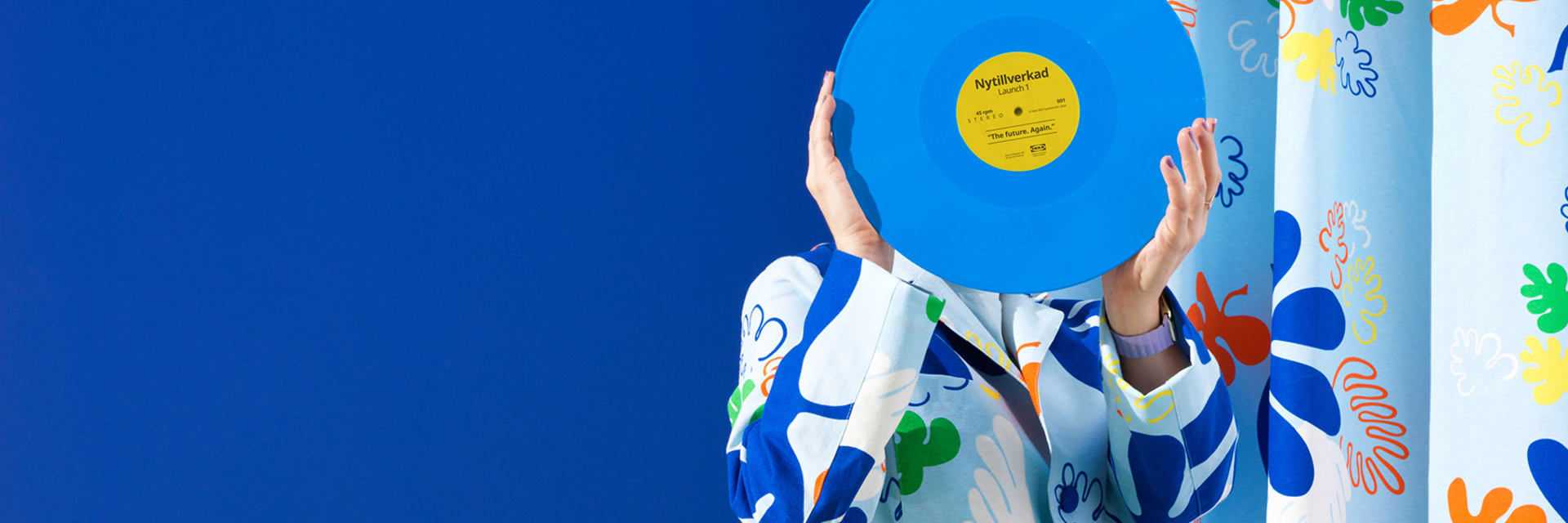 A person stands holding a light blue record in front of their face in a room with a dark blue wall and a blue and orange floral curtain to the right.