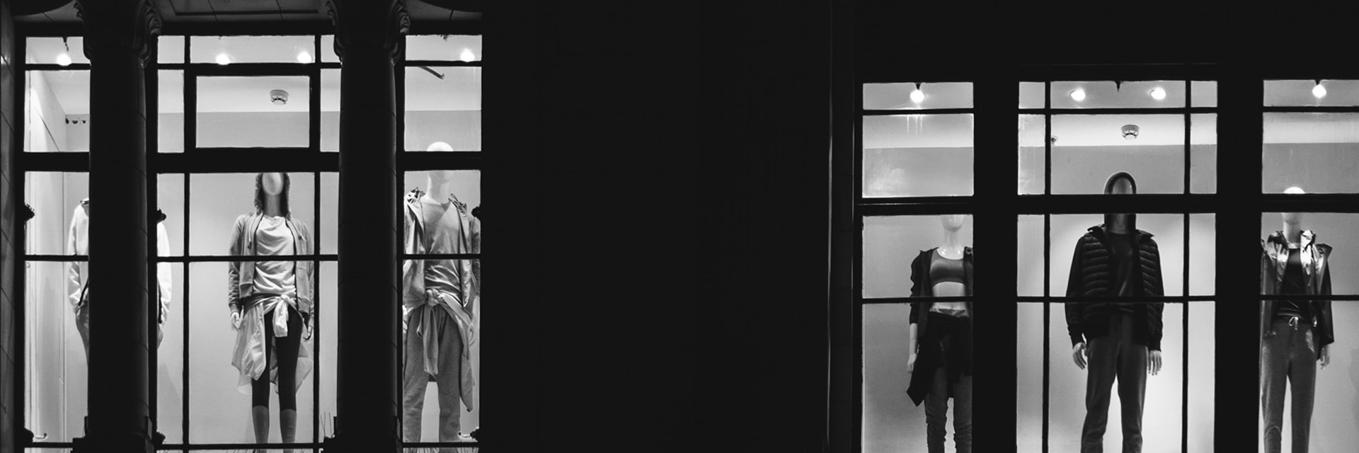 Two sets of windows with mannequins pictured in the dark. On the left and right, three panes show one mannequin each, separated by a black wall. Image taken from outside.