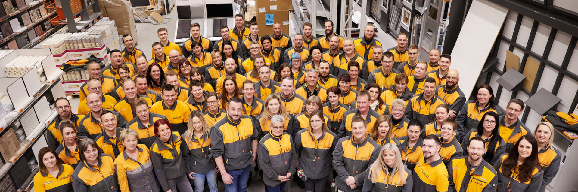 An aerial image taken from ceiling height, of a large group of people smiling up at the camera wearing yellow vests.
