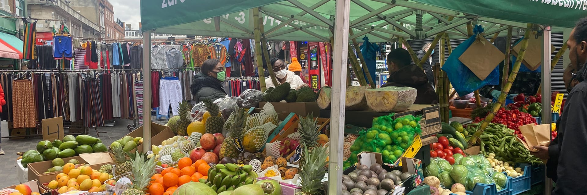 A green awning that reads "Zaro's Fruits and Veg" stands over a large, overflowing fruit stand with three people standing and shopping beneath it in the background.