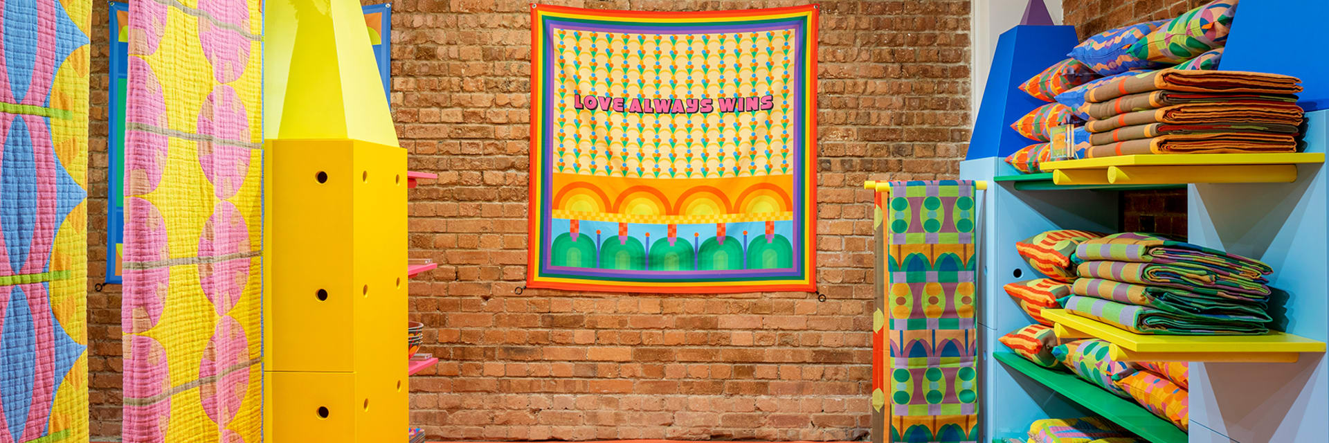 A bright retail space, with yellow shelving and quilted tapestries on the left, green and blue shelving on the right, orange flooring, and a bring wall with a tapestry hanging in the back that reads "Love always wins"
