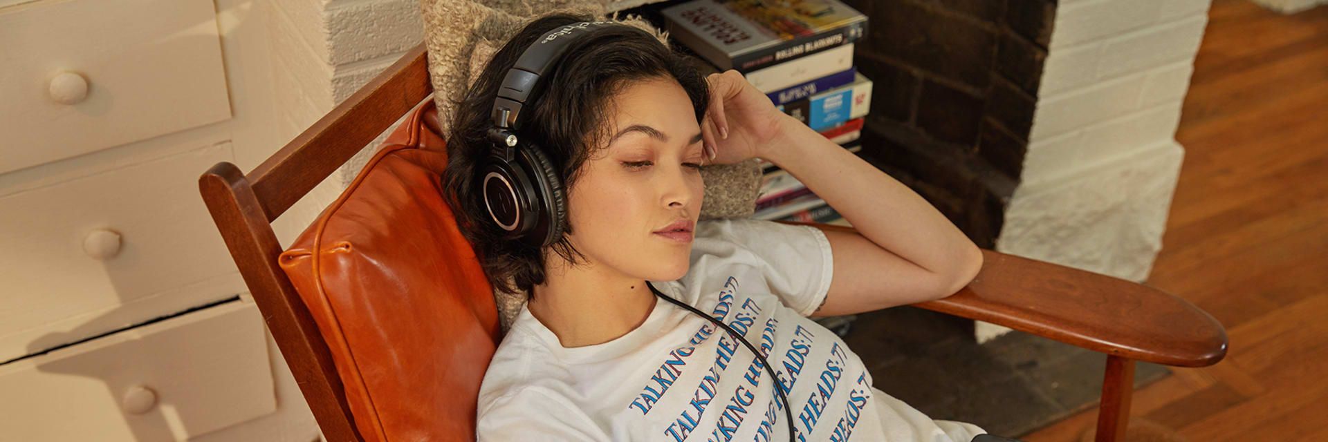 A woman with short dark hair wearing a white t-shirt sits slouched on an armchair with headphones on.