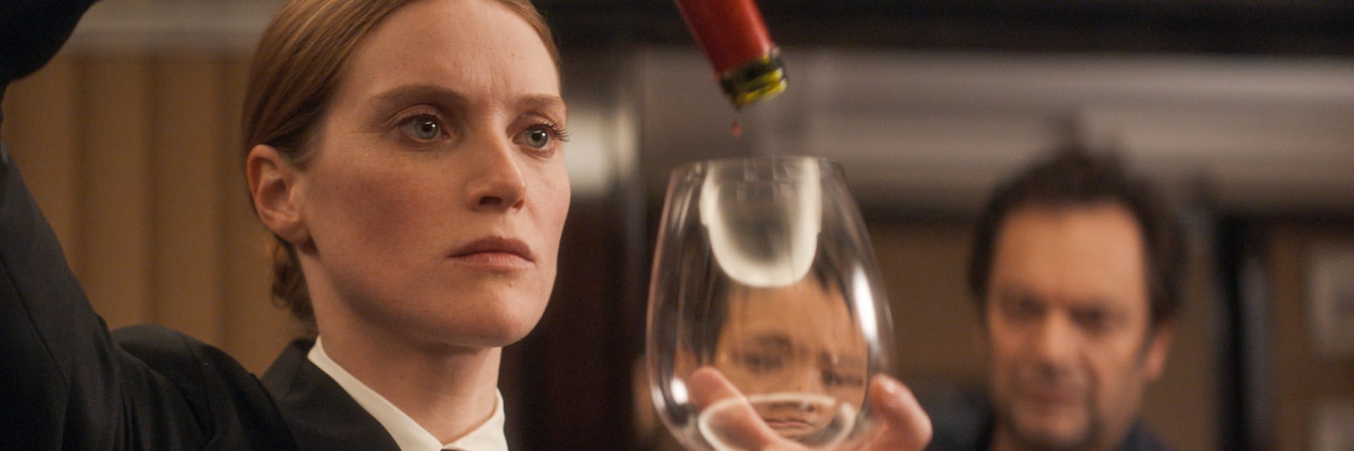 A woman with fair skin and light red hair looks solemly at a wine glass as she pours liquid into it. A man with dark hair watches in the background.