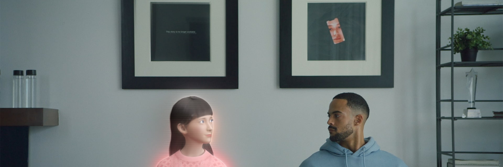 A digitized avatar woman with light skin and dark hair sits on a couch and looks at a real-life man with dark skin and dark hair. He wears a blue sweatshirt.