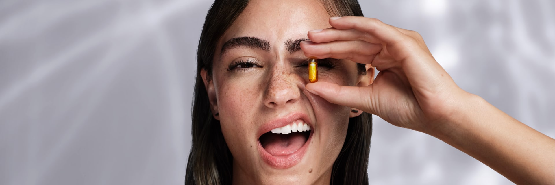 White woman with brown hair and freckles is winking and holding a yellow capsule in front of her left eye She wears a top with thin yellow crisscross strap