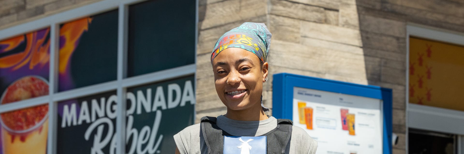 Young black woman in colorful headscarf teeshirt and denim shorts wears a yellow high vis cooling vest Shes standing outside a Dutch Bros store
