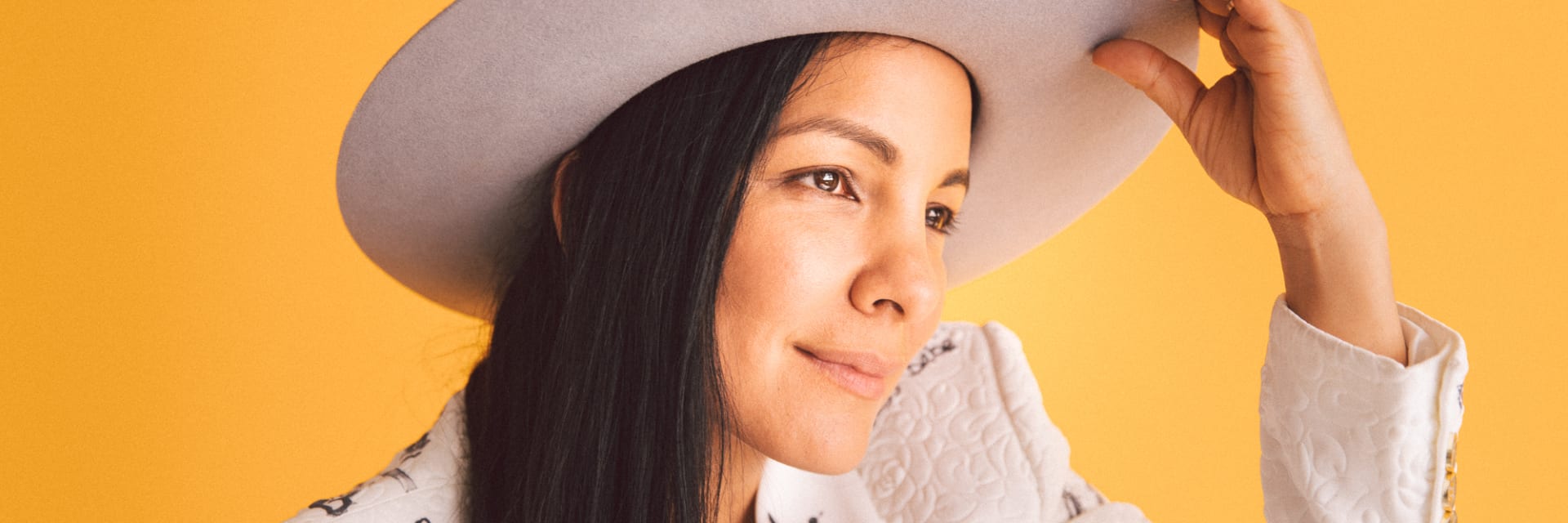A woman with long dark hair wearing a white jacket covered in graffiti and a grey broad brimmed hat smiles looking off camera