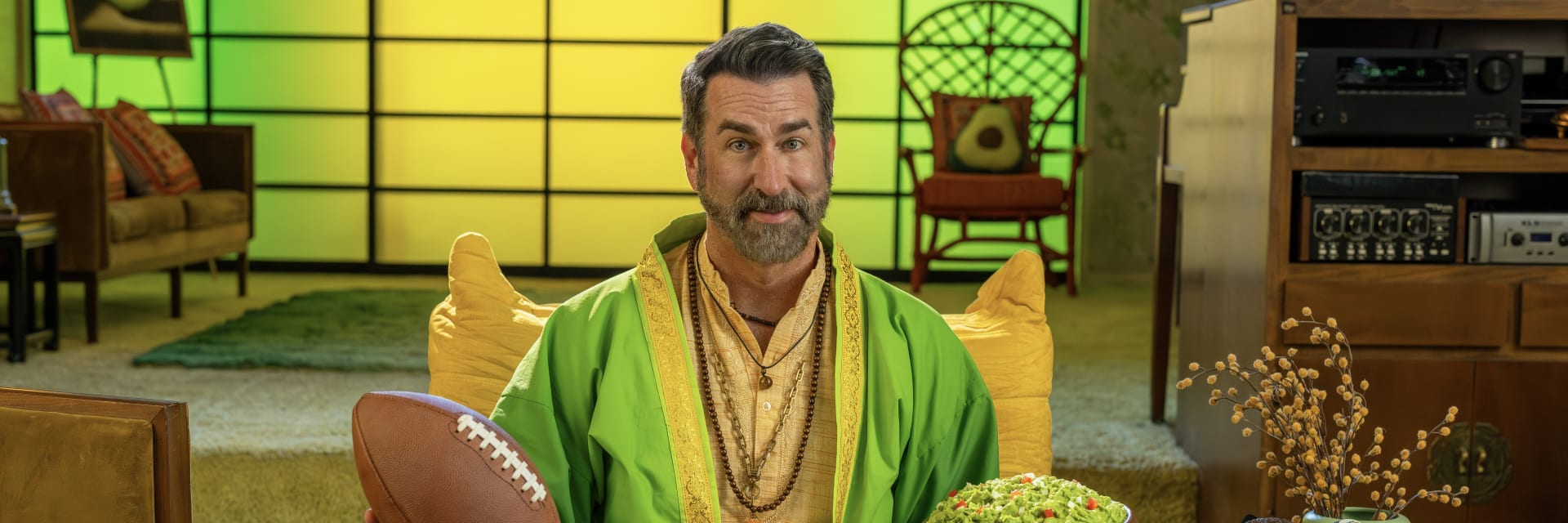 Rob Riggle is sitting cross-legged on a chair, holding a football in one hand and a bowl of guacamole in the other.