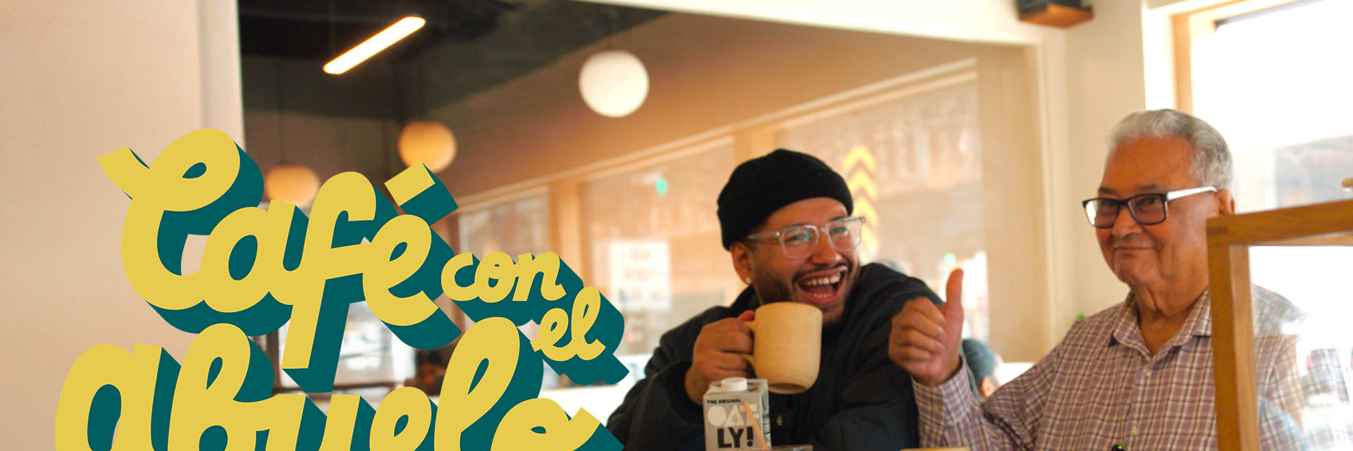 Two Latino men sit at a counter drinking coffee. One is a smiling elder man with white hair and glasses who is giving a thumbsup. The other is a younger man in a black beanie and glasses who is laughing.