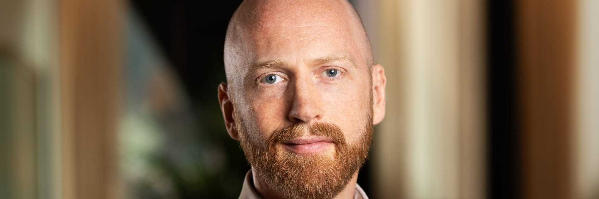 Mikael Kågebäck wears a light button-up shirt and sits against a softly blurred indoor background. He has a shaved head with a reddish-brown beard.
