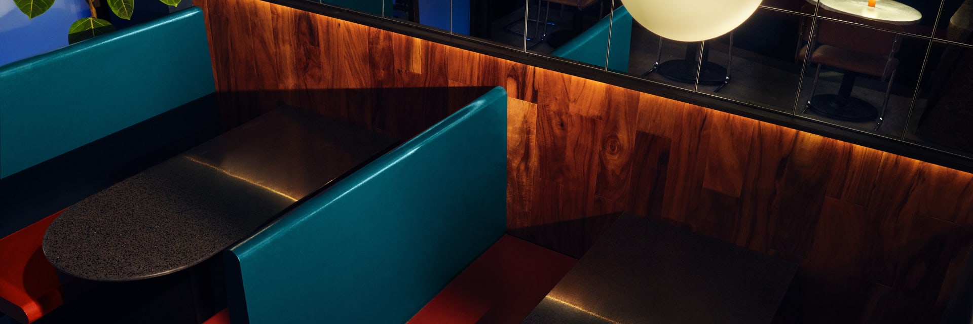 Bar interior featuring paneled wood booths, mirrored wall, teal blue divider screens and round black tables, with retro style globe lamps.
