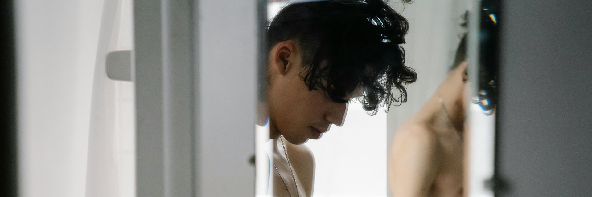 Un joven, de apariencia masculina, con cabello oscuro y rizado con un corte rapado, es capturado en un momento cándido, mirando intensamente un teléfono inteligente que sostiene con ambas manos. Está sin camisa, revelando la parte superior de su cuerpo tonificada, y lleva una cadena plateada con un colgante de cruz. Una pulsera de hilo rojo adorna una de sus muñecas. La imagen está encuadrada como si se viera a través de una puerta o partición ligeramente abierta, con el primer plano borroso en blanco, creando una sensación de intimidad y observación. Al fondo, un espejo refleja parte de su torso desnudo, sugiriendo un ambiente de baño. La iluminación es suave y natural, iluminando el rostro y las manos del sujeto, destacando un momento de concentración tranquila o de interacción con el mundo digital. El estilo general es cándido y personal, con un toque de fotografía naturalista.