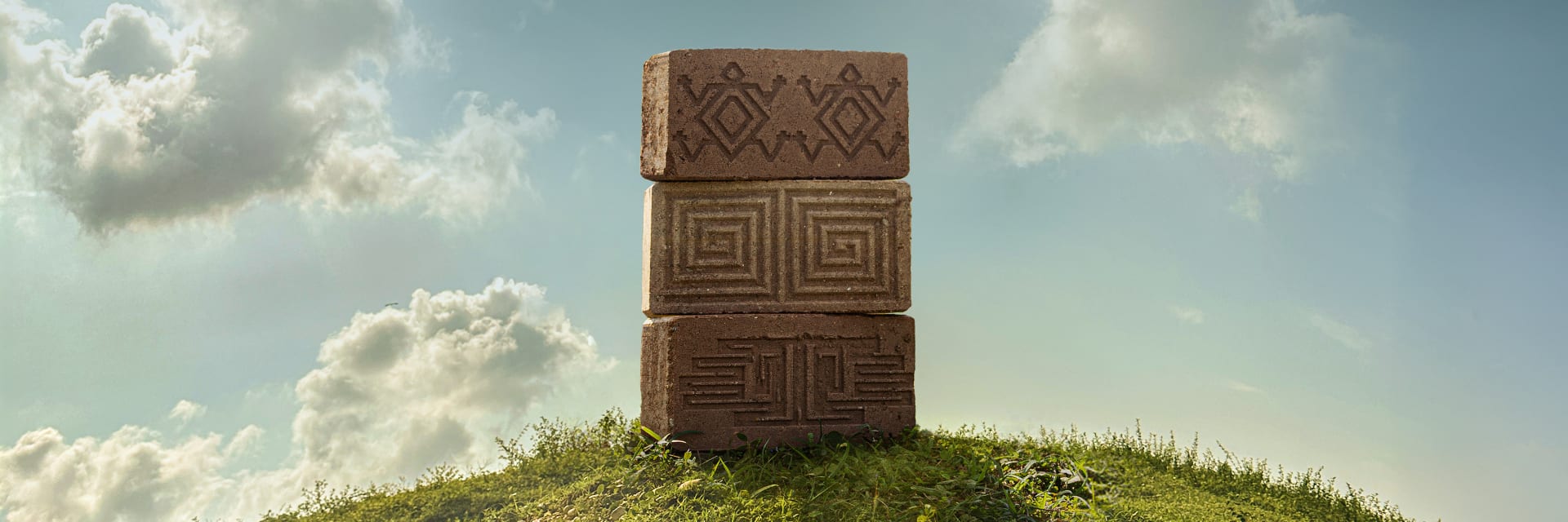 Stacked, textured bricks displaying unique cultural designs sit atop a green hill against a vast sky.
