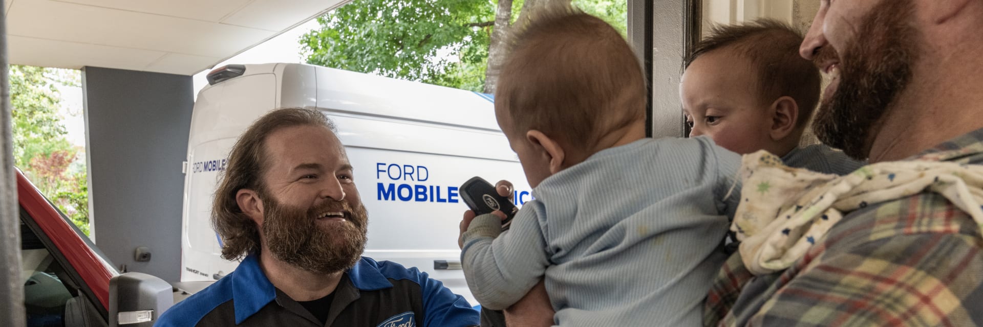 Image of a family standing in the doorway of their home. A dad is holding two babies, one of which is holding a set of keys, and Ford Mobile Services is parked in the driveway.