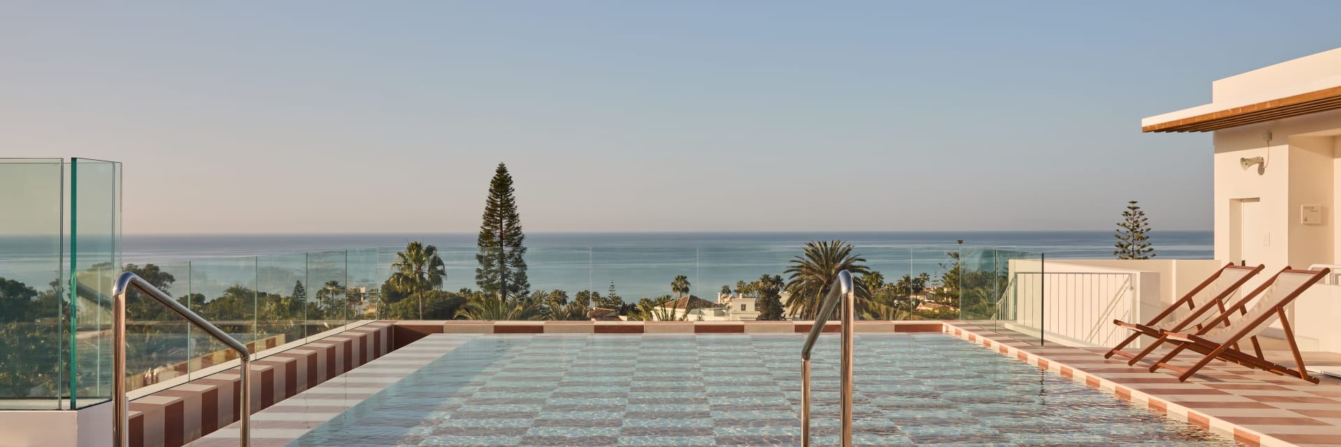 A rooftop terrace featuring a square swimming pool with checkered tiling. The pool deck is made of matching brown and beige square tiles, furnished with a wooden side table, a lounge chair, and sun loungers. In the background, green trees meet the blue ocean under a clear sky.