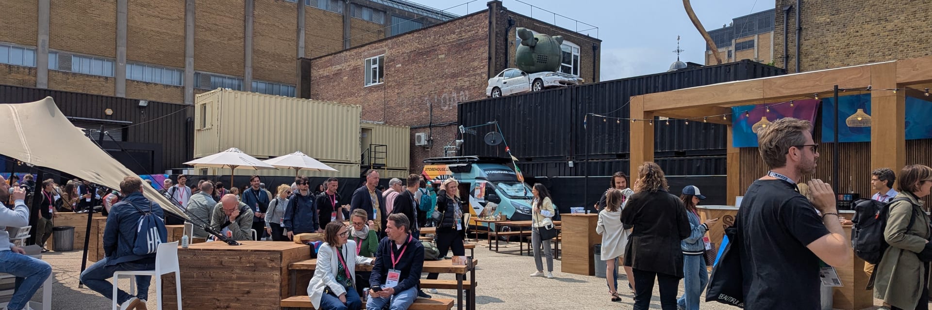 An outdoor, paved courtyard area filled with people socializing in front of industrial-style buildings. Some people are sitting at wooden picnic tables and high-top bar stools under beige umbrellas, while others walk through the space. In the background, there are shipping containers, brick buildings, a white car parked on a lower rooftop, and a large, stylized bow sculpture pointing toward the sky on the right.