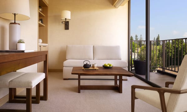 Image of a cream-colored luxury suite with a high table, coffee table, a loveseat and an armchair. The room sits adjacent to sliding doors and a balcony with scenic views.