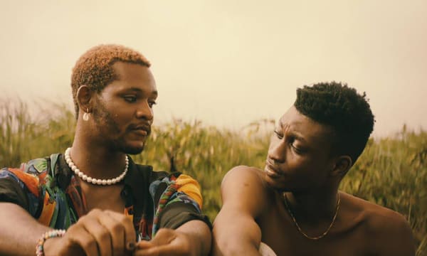 Two Black men sitting in the grass, exchanging a tender glance