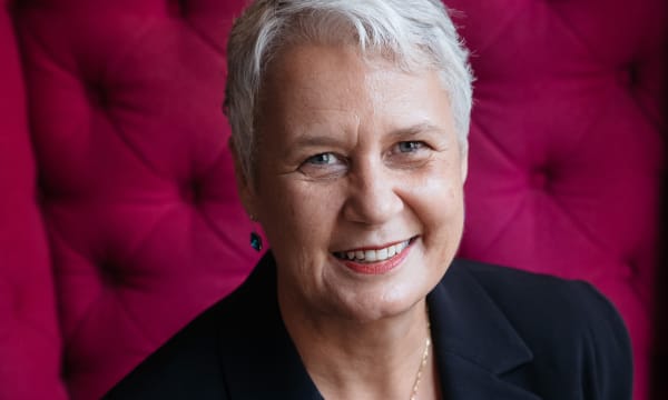 A white, older woman with short white hair smiles off-center at the camera. She wears a black shirt and blue gemstone earrings in front of a red background.