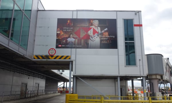 OOH Billboard at airport which features HSBC logo and an older lady playing guitar and text: 'What can we learn from teaching others?'
